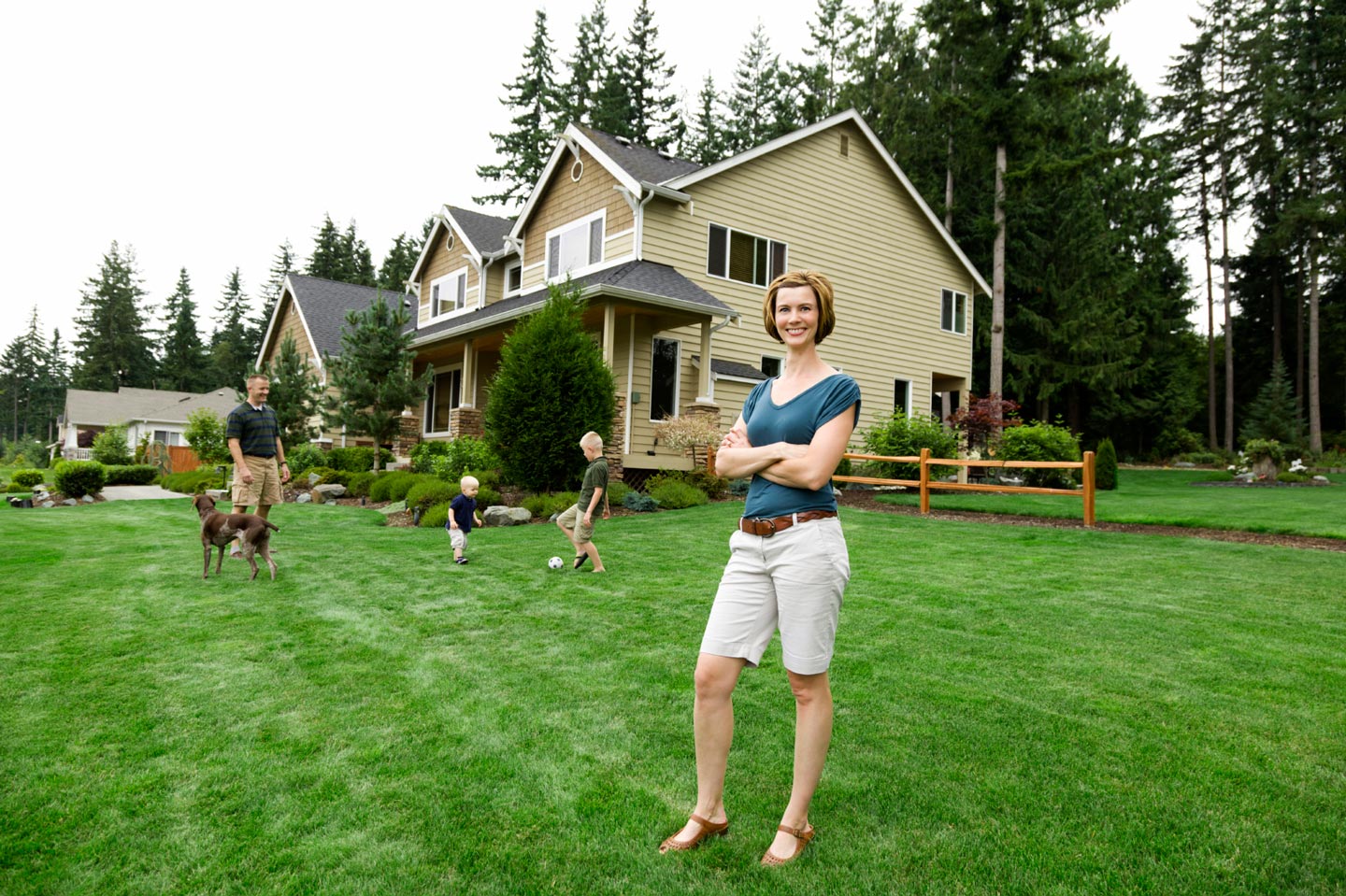 A mother smiles confidently as her children play with her husband and the family dog in the yard in front of their home.