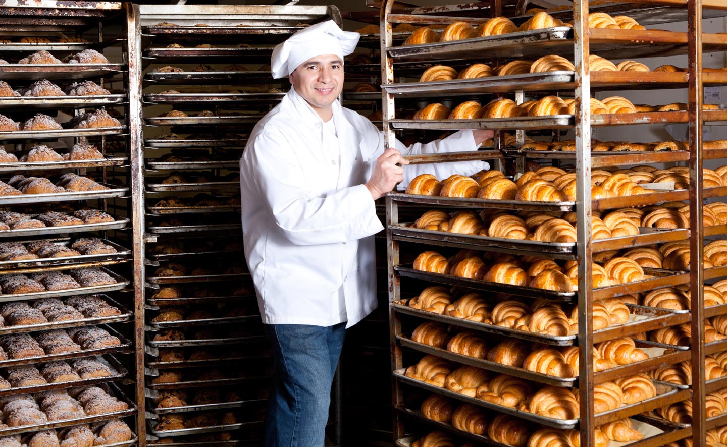 A baker stands next to one of many racks filled with baked goods.